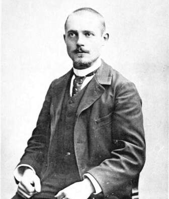Charles Péguy, seated, looking upward with shaved head, moustache, and goatee, dressed in a full suit with round white collar.