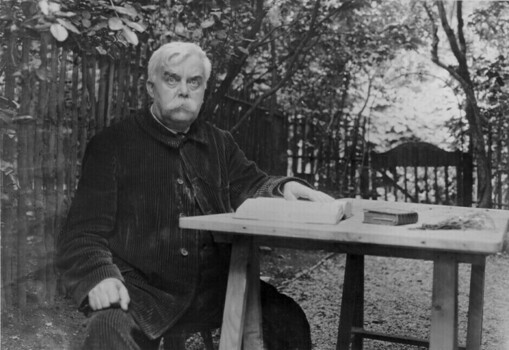 Léon Bloy, seated with books in garden, wearing a corduroy overcoat, with big white moustache, staring directly toward the camera.