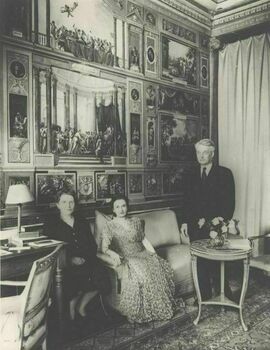 Jacques standing in a dark suit, Raïssa and Vera seated beside him, with a wall full of classical mural paintings behind them.