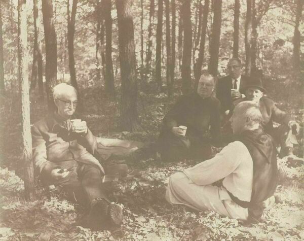 Jacques Maritain seated on the forest floor, drinking coffee and smoking a pipe, Thomas Merton, wearing a habit, seated with his back towards the camera and looking at Maritain with three other onlookers.