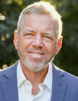 man with gray hair and beard, wearing blue jacket and white oxford, smiling before trees and evening sunlight