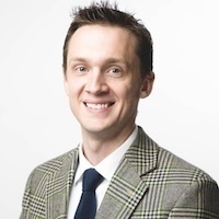A smiling man with light brown hair wears a gray and green plaid jacket, white shirt, and navy blue tie.