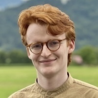 Young man with red hair and glasses, wearing a tan shirt, smiles outdoors with a green field and hills behind him.